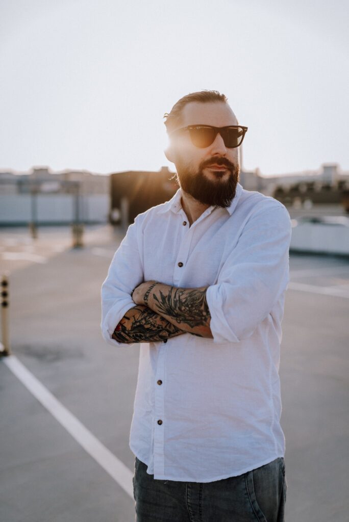 mężczyzna z brodą w okularach przeciwsłonecznych, w białej koszuli i z tatuażami na rękach na tle zachodzącego słońca