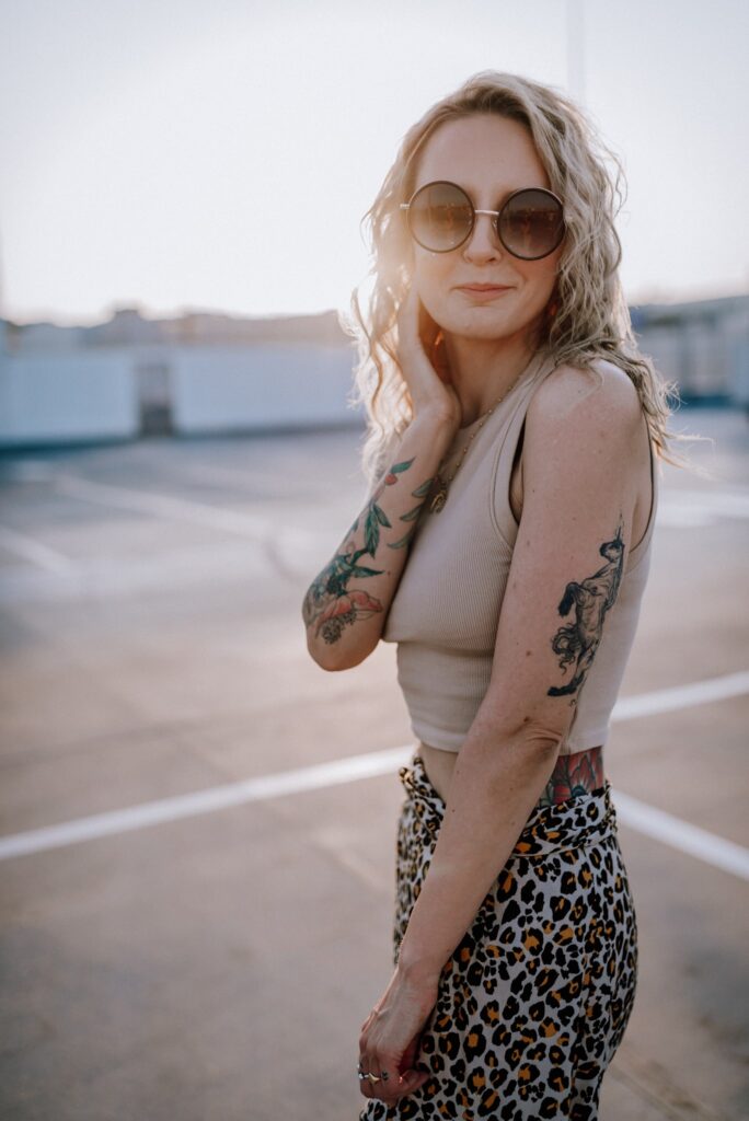 dziewczyna z tatuażami w okrągłych okularach przeciwsłonecznych, bezowym topie i spódnicy w panterkę na tle zachodzącego słońca