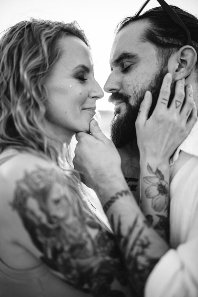 Zakochana para w bliskim ujęciu na czarno-białym zdjęciu. Kobieta delikatnie dotyka twarzy mężczyzny, ich czoła są złączone, a on uśmiecha się do niej czule. Oboje mają liczne tatuaże, które są widoczne na ich ramionach i dłoniach. Tło jest rozmyte, skupiając uwagę na ich wzajemnym spojrzeniu i emocjach