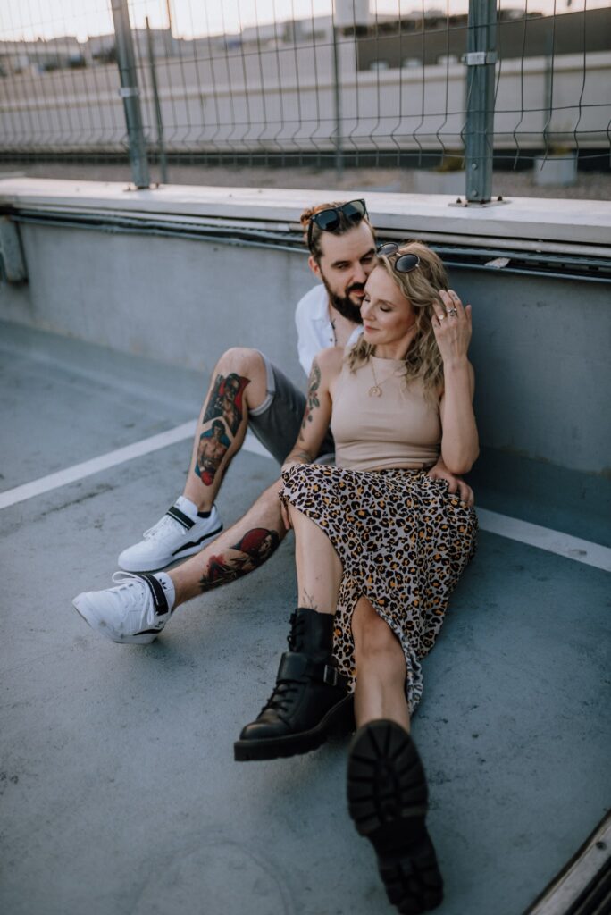 para siedząca na ziemi i przytulająca się, czule spogladajać na siebie i smiejąc się, ona ma zadartą nogę do góry na parkingu na tle barierek