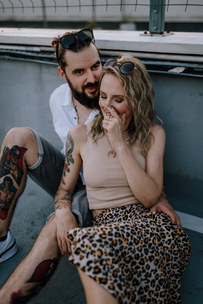 para siedząca na ziemi i przytulająca się, czule spoglądając na siebie i śmiejąc się na parkingu na tle ściany