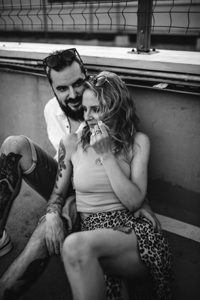 para siedząca na ziemi i przytulająca się, czule spoglądając na siebie i śmiejąc się na parkingu na tle ściany, zdjecie czarno białe