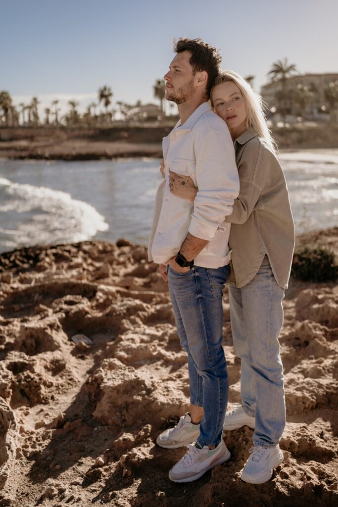 kobieta przytula się do pleców mężczyzny stojąc na skałach nad morzem.