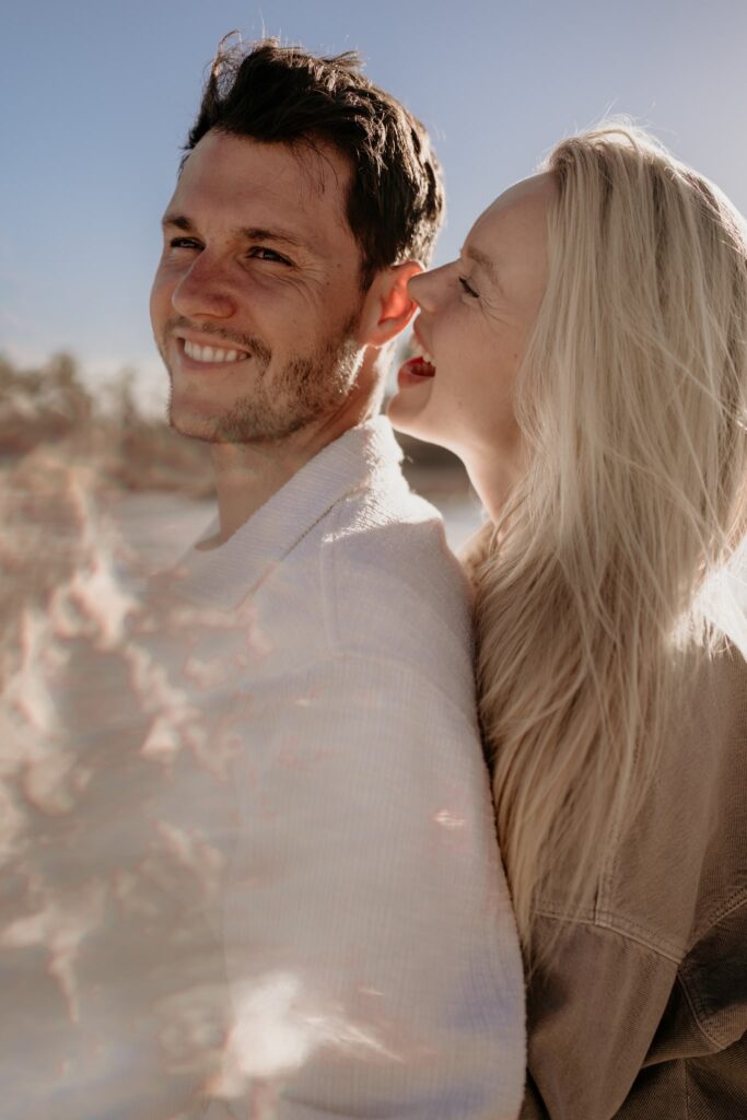 Para w miłosnym uścisku na plaży, kobieta śmieje się, szepcząc coś do ucha partnera, sesja zdjęciowa o zachodzie słońca.