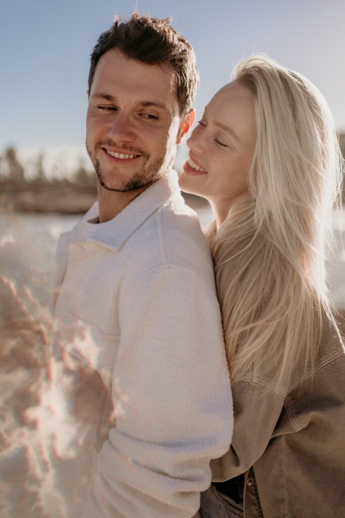 Para w miłosnym uścisku na plaży, kobieta śmieje się, szepcząc coś do ucha partnera, sesja zdjęciowa o zachodzie słońca.