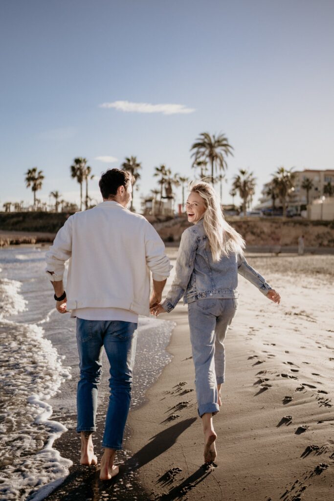 Para trzymająca się za ręce, biegająca boso po plaży w Torrevieja podczas złotej godziny. Kobieta patrzy na mężczyznę z uśmiechem, a za nimi widać morskie fale i palmy.