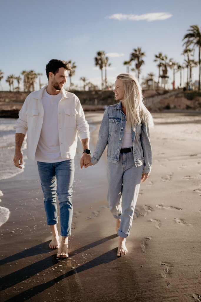 Para trzymająca się za ręce, spacerująca boso po plaży w Torrevieja podczas złotej godziny. Kobieta patrzy na mężczyznę z uśmiechem, a za nimi widać morskie fale i palmy.