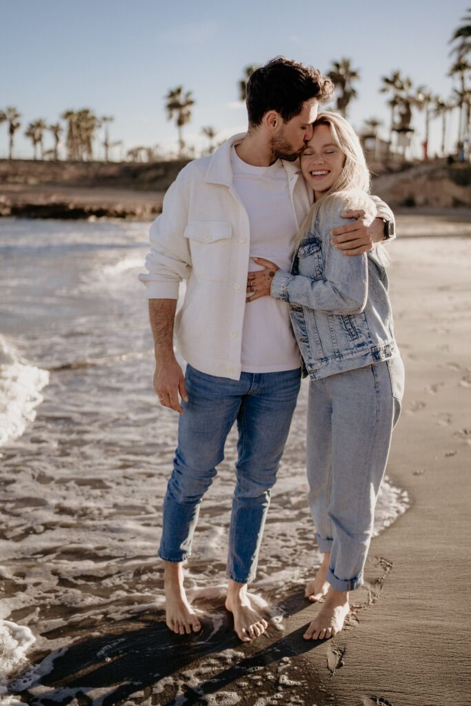 Para na plaży w Torrevieja, kobieta z długimi blond włosami, ubrana w jeansową kurtkę, uśmiecha się szeroko, gdy mężczyzna całuje ją w czoło. W tle palmy i morze o złotej godzinie.