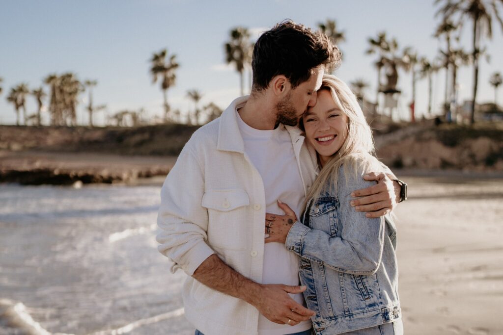 Para na plaży w Torrevieja, kobieta z długimi blond włosami, ubrana w jeansową kurtkę, uśmiecha się szeroko, gdy mężczyzna całuje ją w czoło. W tle palmy i morze o złotej godzinie.