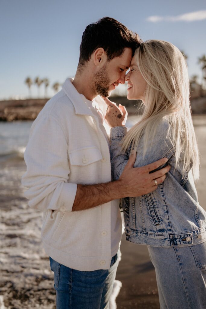 Zakochana para zbliżająca się do siebie z czułym uśmiechem podczas złotej godziny, sesja narzeczeńska na plaży.