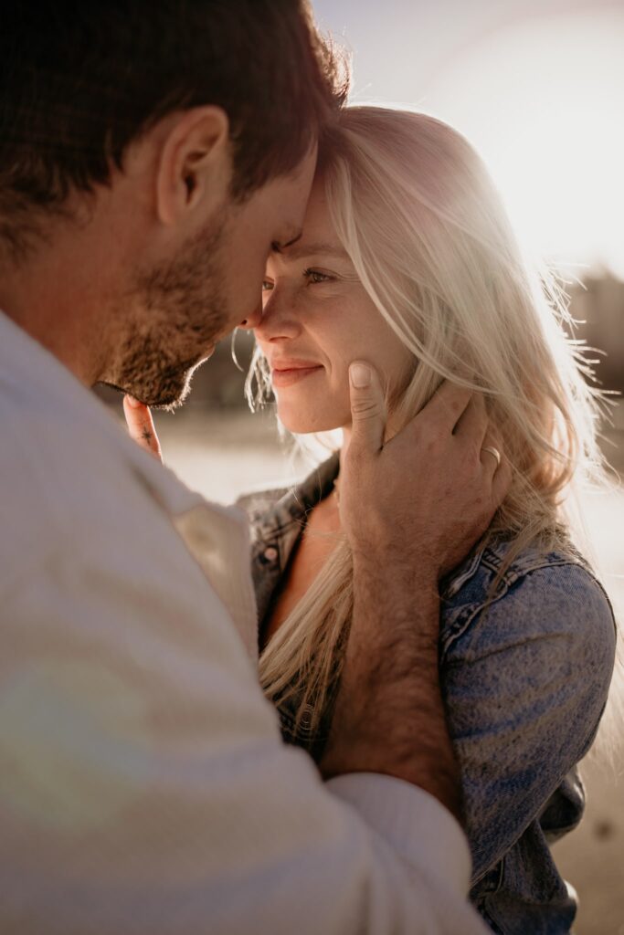 Zakochana para zbliżająca się do siebie z czułym uśmiechem podczas złotej godziny, sesja narzeczeńska na plaży.