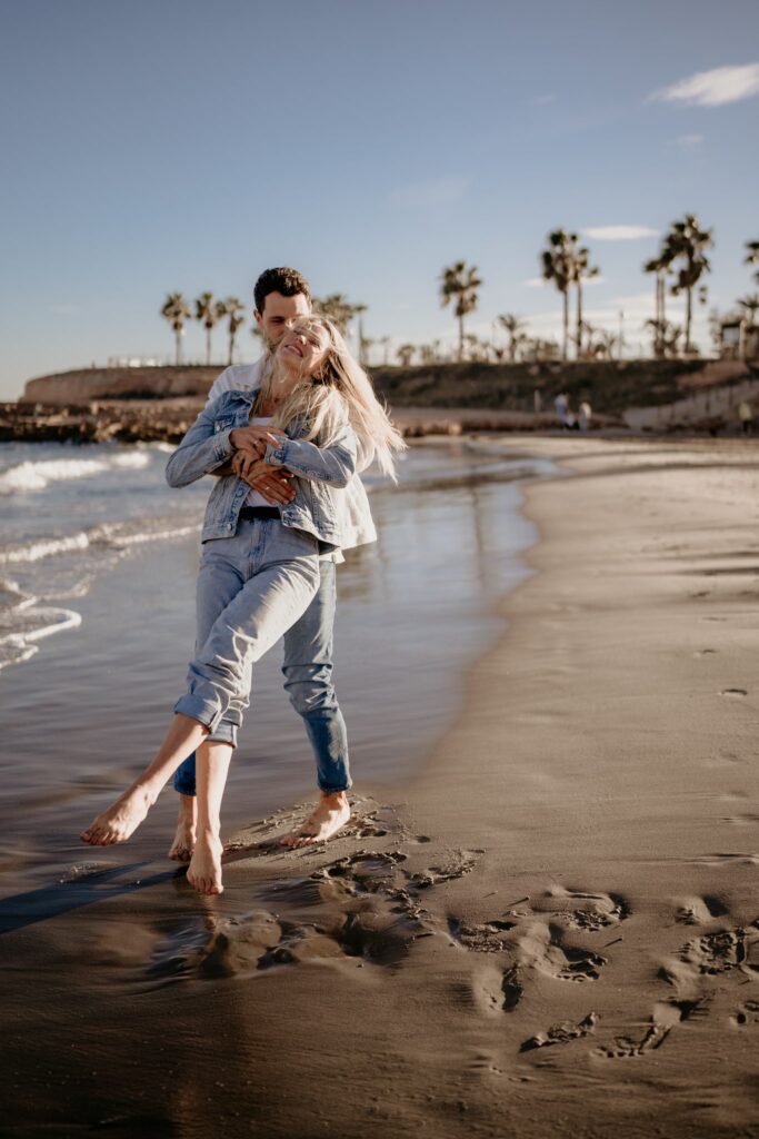 Para na plaży w Torrevieja, kobieta z długimi blond włosami, ubrana w jeansową kurtkę, uśmiecha się szeroko, gdy mężczyzna obejmuje ją od tyłu i unosi. W tle palmy i morze o złotej godzinie.
