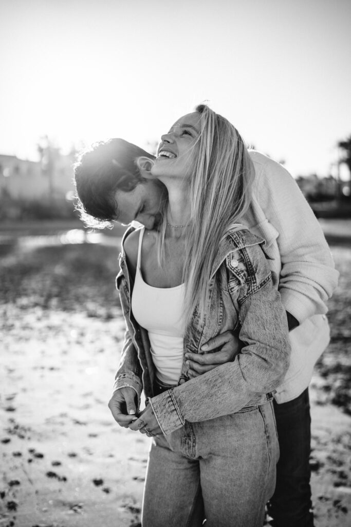 Mężczyzna całuje kobietę w szyję, trzymając ją w objęciach na plaży w Torrevieja. Oboje są ubrani na luzie, w jeansy i jasne kurtki, a kobieta śmieje się, spoglądając na w gorę zdjecie czaro białe