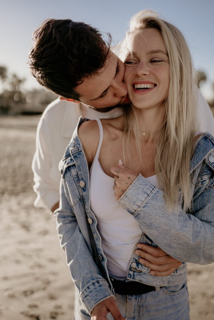 Mężczyzna całuje kobietę w szyję, trzymając ją w objęciach na plaży w Torrevieja. Oboje są ubrani na luzie, w jeansy i jasne kurtki, a kobieta śmieje się, zerkając w jego stronę