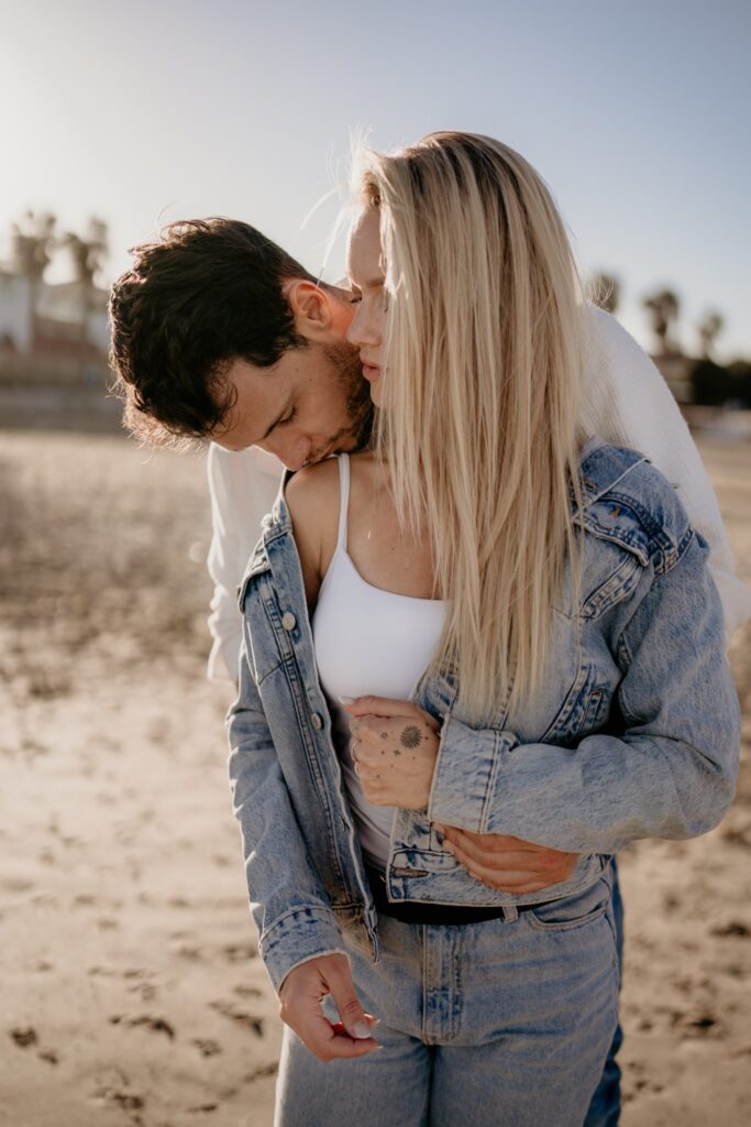Mężczyzna całuje kobietę w ramię, trzymając ją w objęciach na plaży w Torrevieja. Oboje są ubrani na luzie, w jeansy i jasne kurtki, a kobieta patrzy w jego stronę