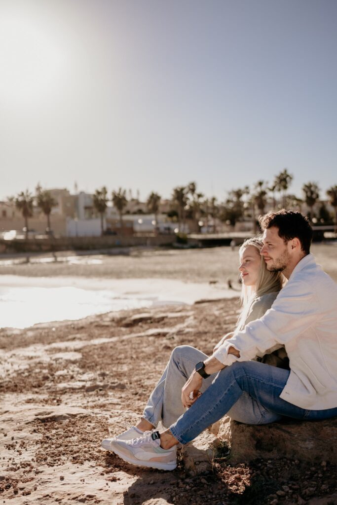 para siedząca na skałach nad morzem, kobieta opiera sie placami o mężczyzne a aon ją obejmuję, patrza na horyznt